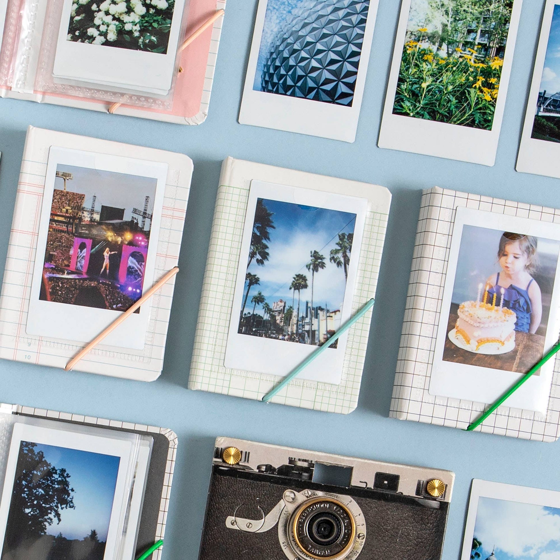 A styled flat-lay arrangement of instant photograph prints, some mounted on notebooks with colored pencils, alongside a vintage camera on a light blue background.
