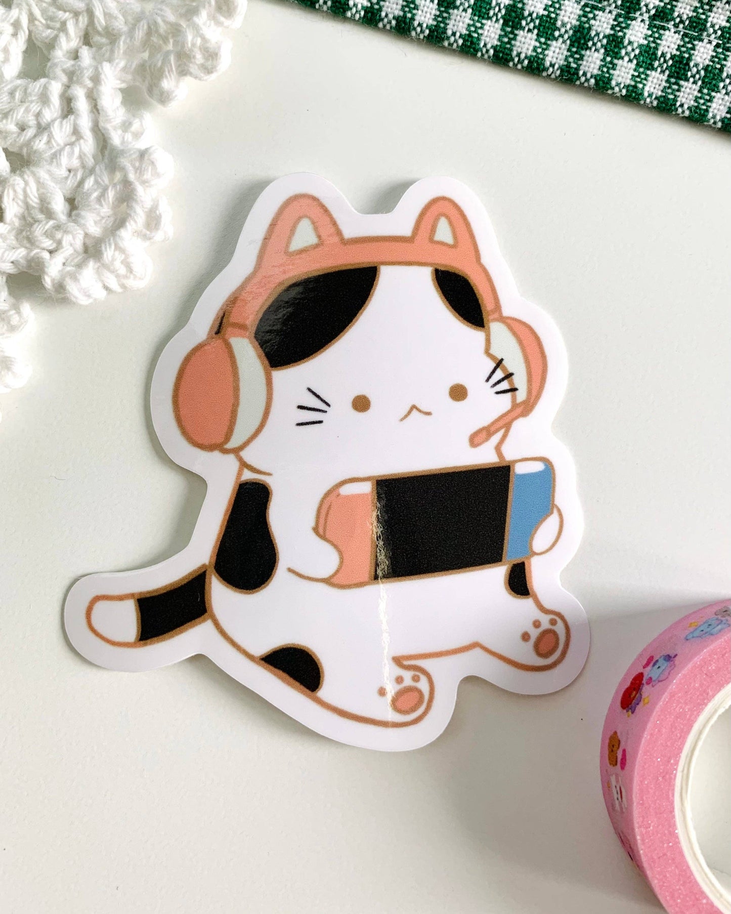 Sticker of a cat with headphones on a white surface with a green checkered cloth in the background. Black and White Gamer Kitty Sticker