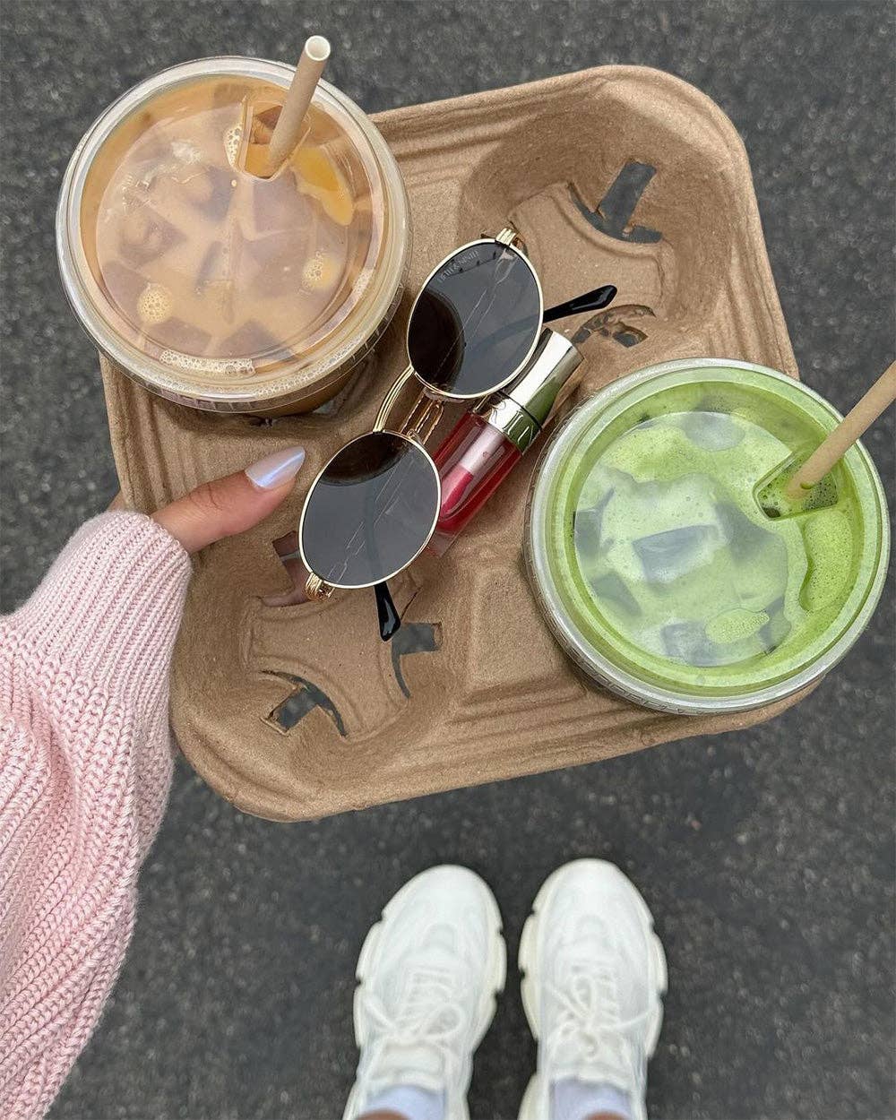 Two drinks in plastic cups with straws, a pair of sunglasses, and a hand holding a pink sleeve on a brown tray.