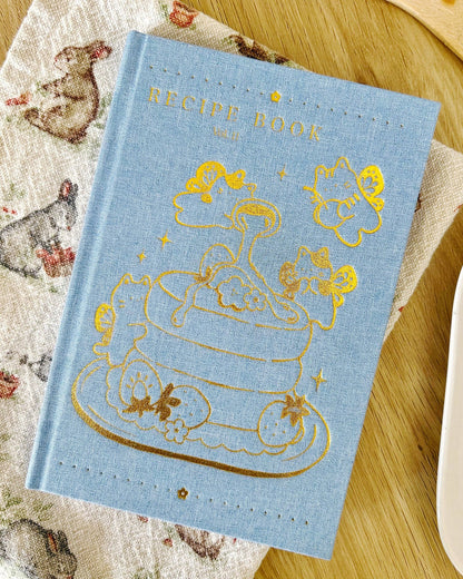 Blue recipe book with gold illustrations on a wooden surface