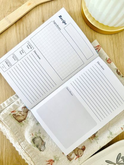 Two recipe cards on a wooden surface with a floral cloth underneath. Blue Linen Recipe Book