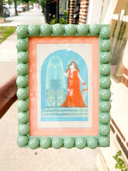 A hand holds a square decorative picture frame outdoors. The frame has a mint green beaded border and a peach inner mat surrounding a vintage-style illustration of a woman in a red dress under a blue arch.