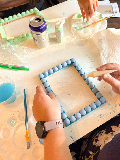 A person's hands are shown painting a square picture frame with light blue beads, working at a white craft table surrounded by paint brushes, a paint palette, and other painting supplies.