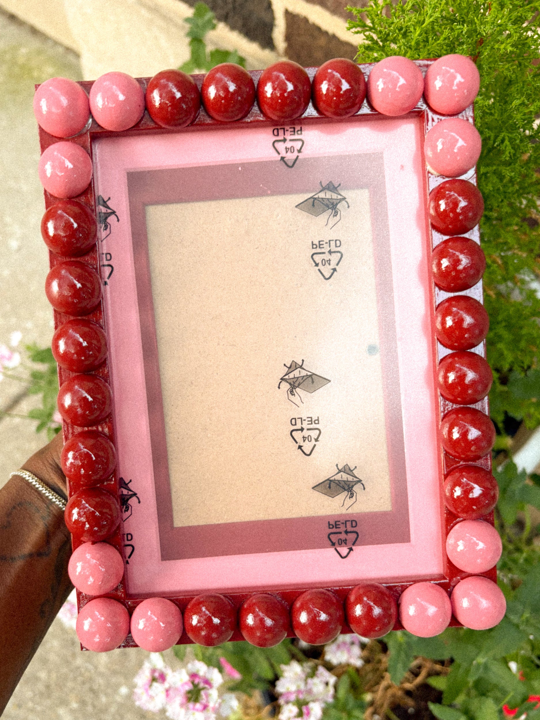 A square decorative picture frame with a pink inner border and a beaded outer border made of glossy round beads in shades of pink and red. The frame is being held outdoors by a hand, with green foliage and flowers in the background.