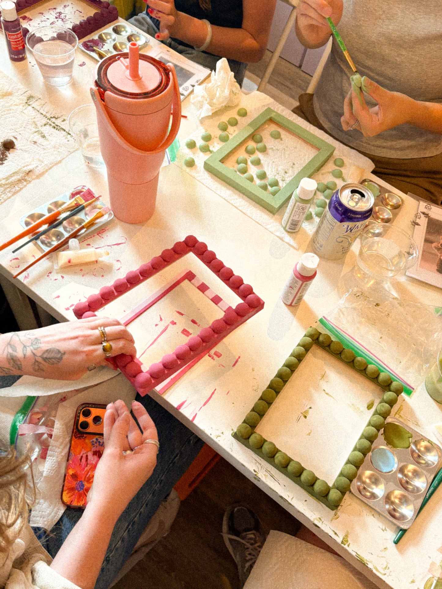 A group of people painting colorful rectangular frames made of small round beads, working at a white table covered with paintbrushes, paint bottles, and art supplies.