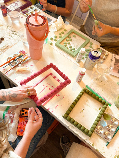 A group of people painting colorful rectangular frames made of small round beads, working at a white table covered with paintbrushes, paint bottles, and art supplies.