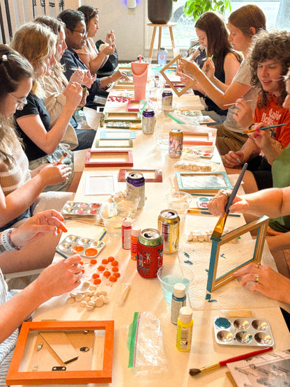 A group of people sitting around a long table, painting and assembling framed art pieces in a bright workshop or studio setting.