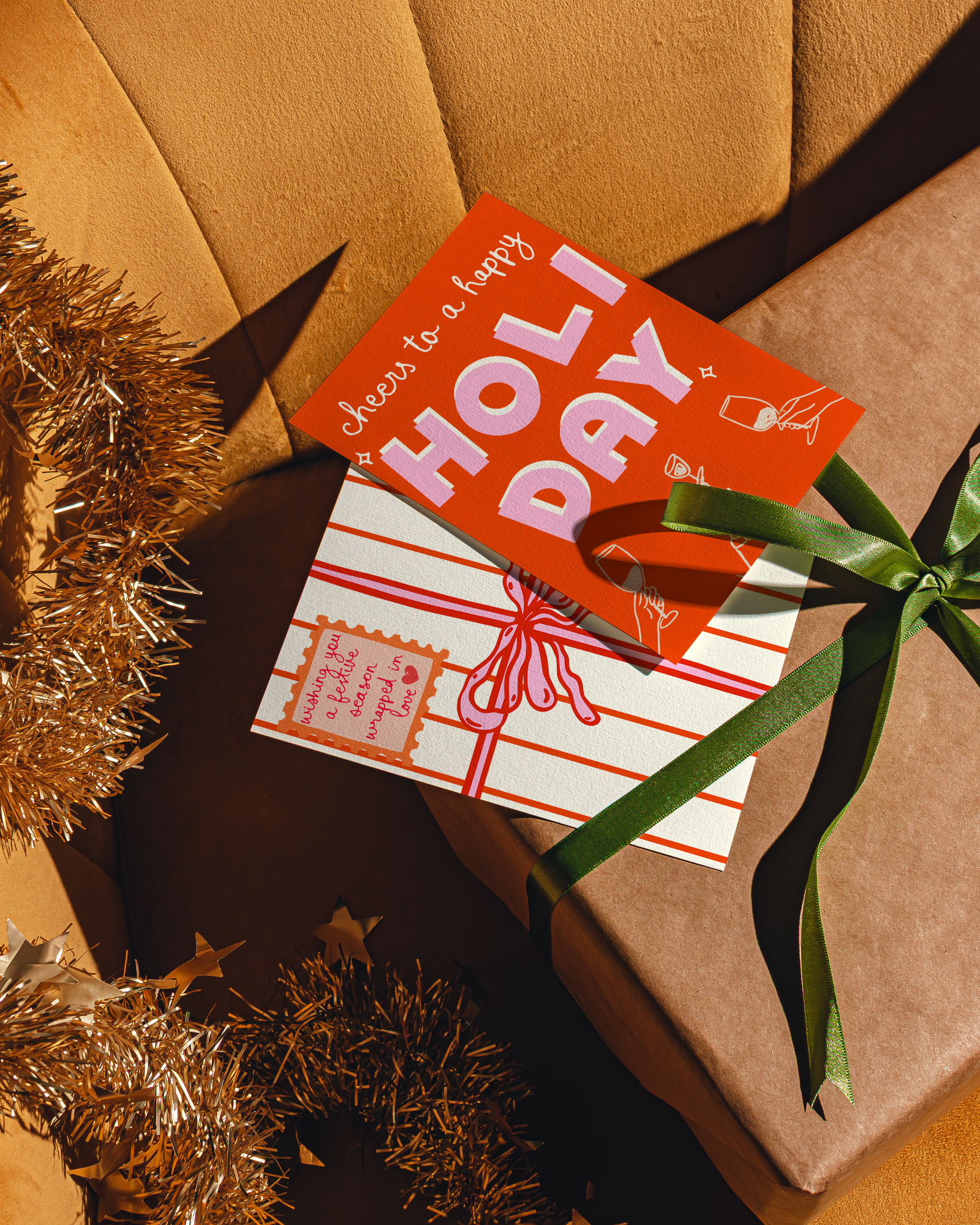 Gift box with a card that says 'Cheers to a happy HOLIDAY' on an orange sofa surface with tinsel.