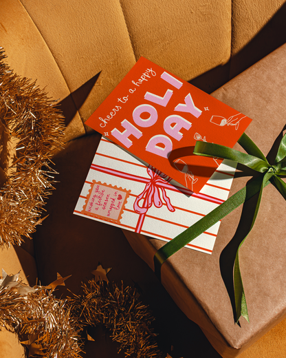 Gift box with a card that says 'Cheers to a happy HOLIDAY' on an orange sofa surface with tinsel.