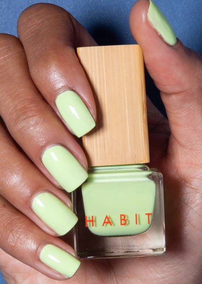 Hand holding a bottle of green nail polish with 'Habit' branding against a blue background