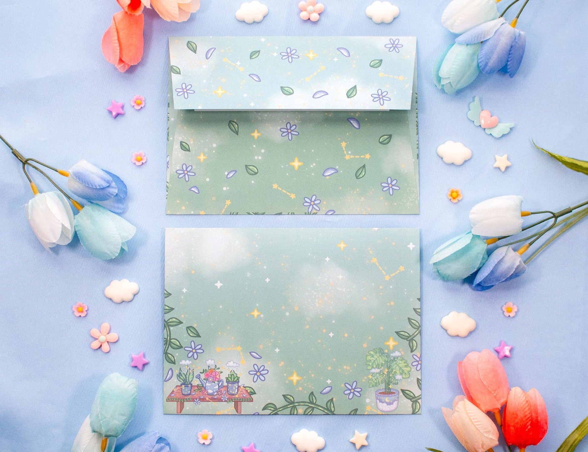 A flat-lay presentation of two pastel green paper envelopes decorated with floral and celestial patterns, surrounded by decorative tulips, stars, and clouds on a light blue background.