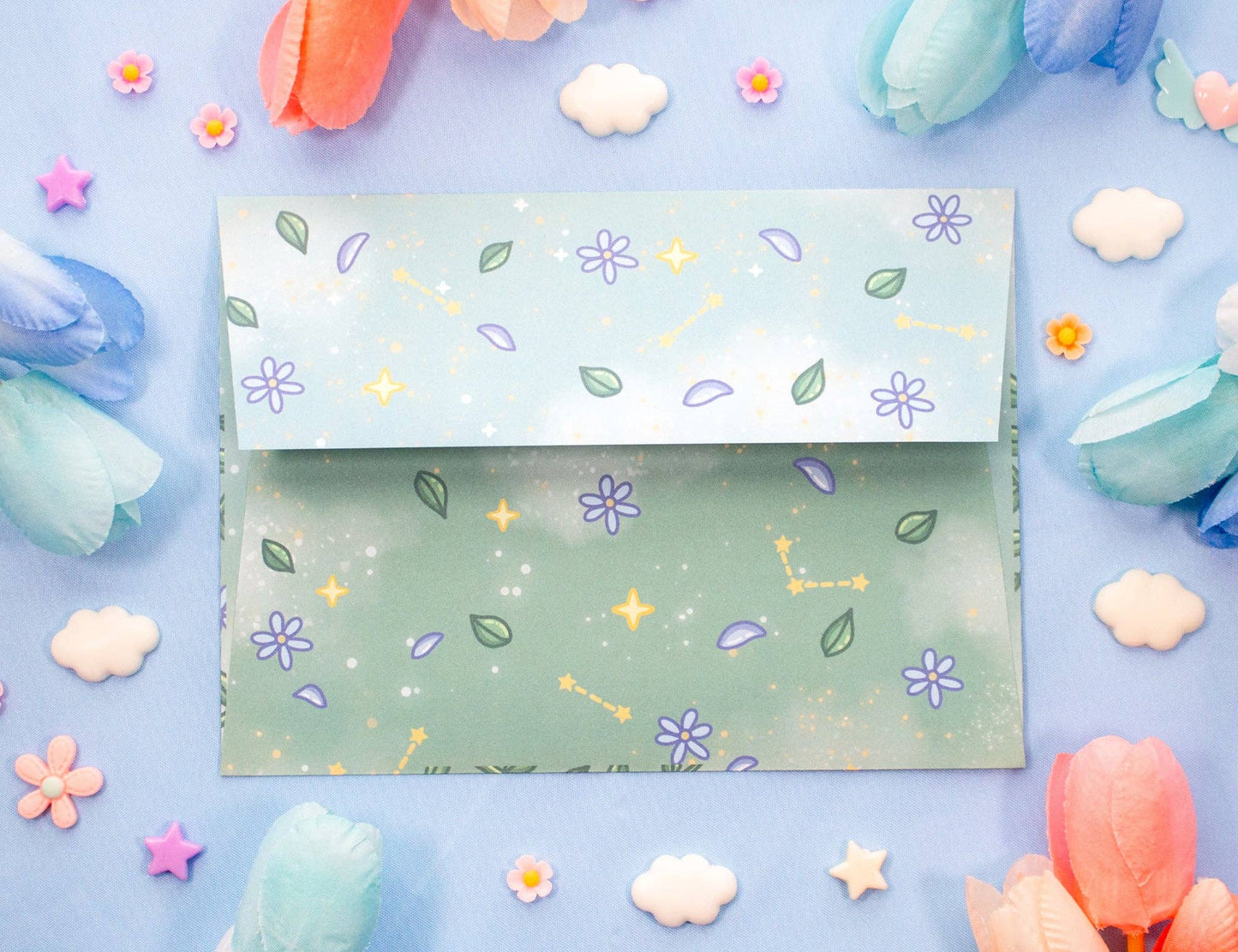 A pastel blue and green patterned envelope with purple flowers, green leaves, and gold stars, displayed on a light blue background surrounded by decorative paper flowers and clouds.