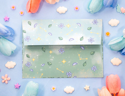 A pastel blue and green patterned envelope with purple flowers, green leaves, and gold stars, displayed on a light blue background surrounded by decorative paper flowers and clouds.