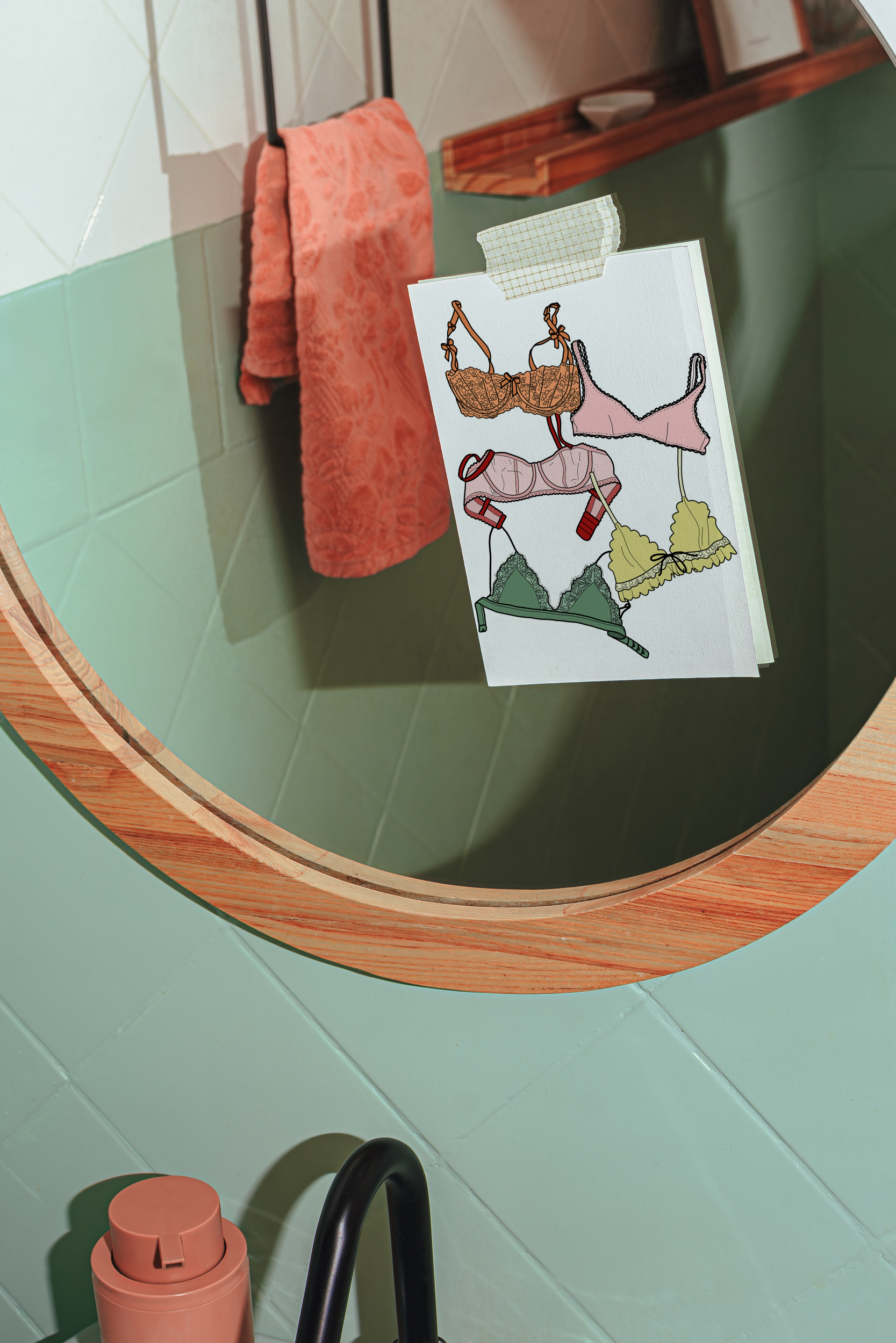 Round mirror with wooden frame on a green wall, featuring a small art print with illustrations of bras in various colors.