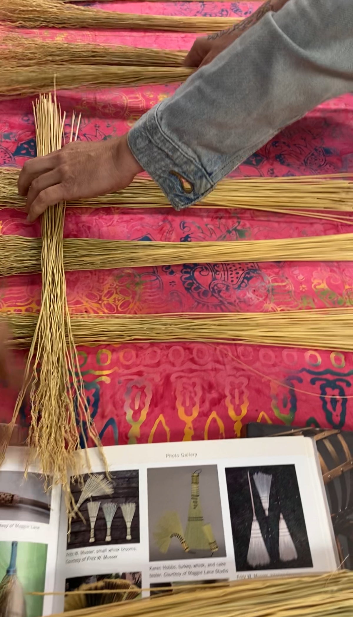 A close-up of hands weaving or arranging long strands of natural fiber on a bright pink patterned fabric, with an open printed booklet visible at the bottom.