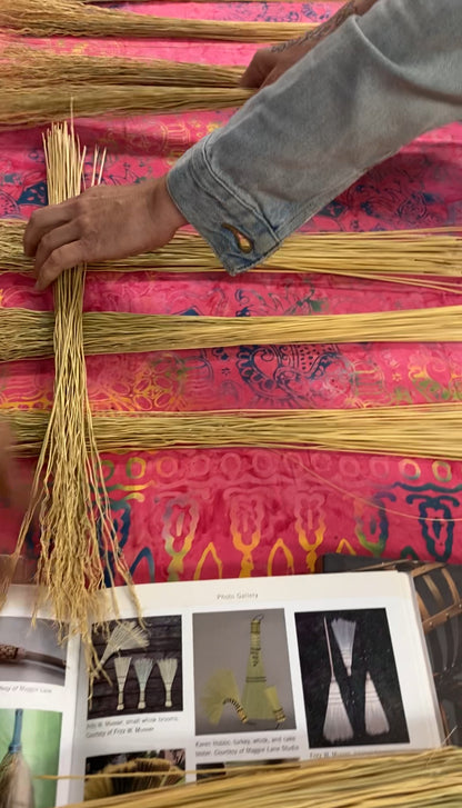 A close-up of hands weaving or arranging long strands of natural fiber on a bright pink patterned fabric, with an open printed booklet visible at the bottom.