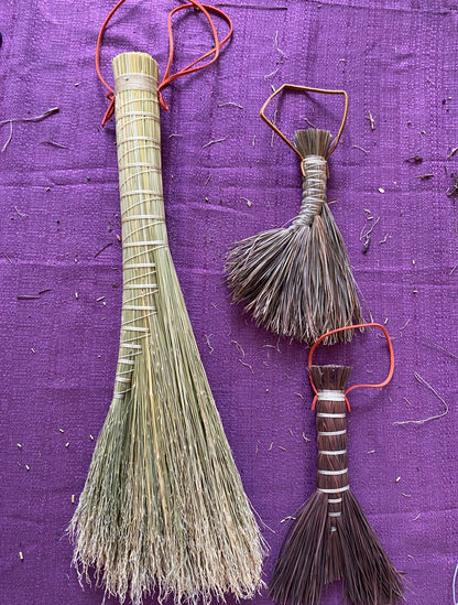 Two traditional hand-woven straw brooms with red hanging loops, displayed on a textured purple fabric background.