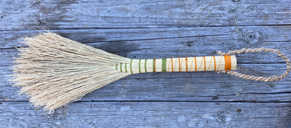 A traditional hand-woven straw broom with a wrapped handle and hanging loop, displayed flat on a weathered blue-gray wooden plank surface.