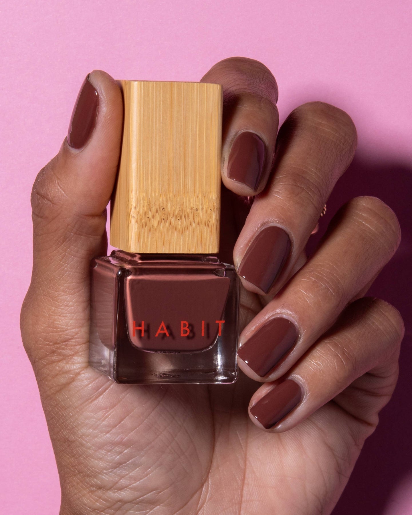 Hand holding a red nail polish bottle with 'Habit' branding against a pink background. Brownie Points Brown Toxin Free Nail Polish