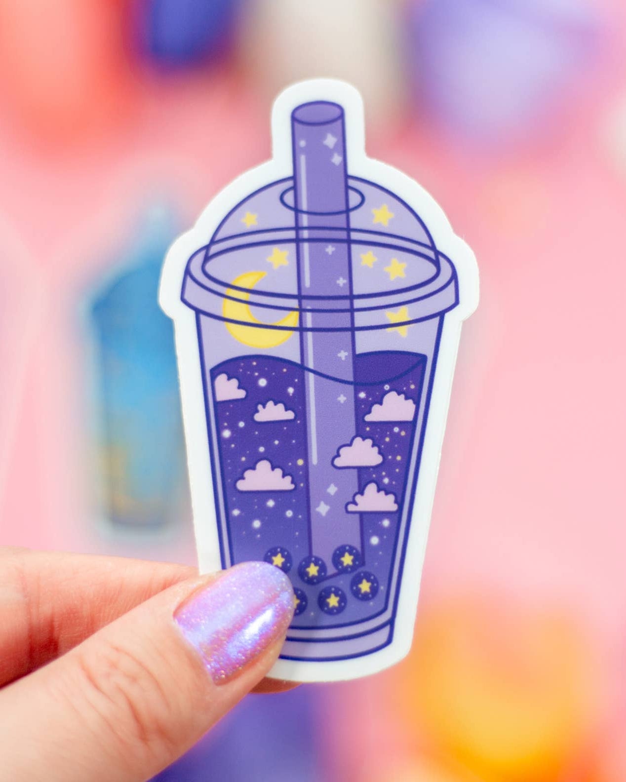 Sticker of a purple bubble tea with clouds and stars held by a hand against a colorful blurred background