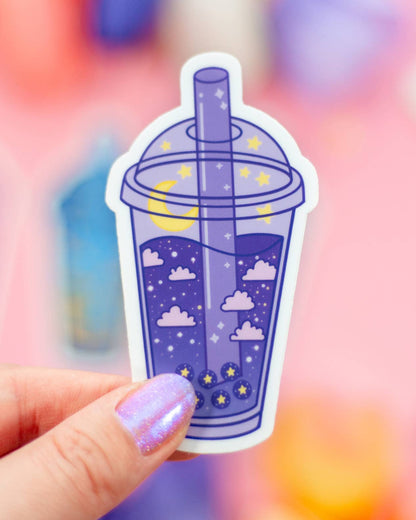 Sticker of a purple bubble tea with clouds and stars held by a hand against a colorful blurred background
