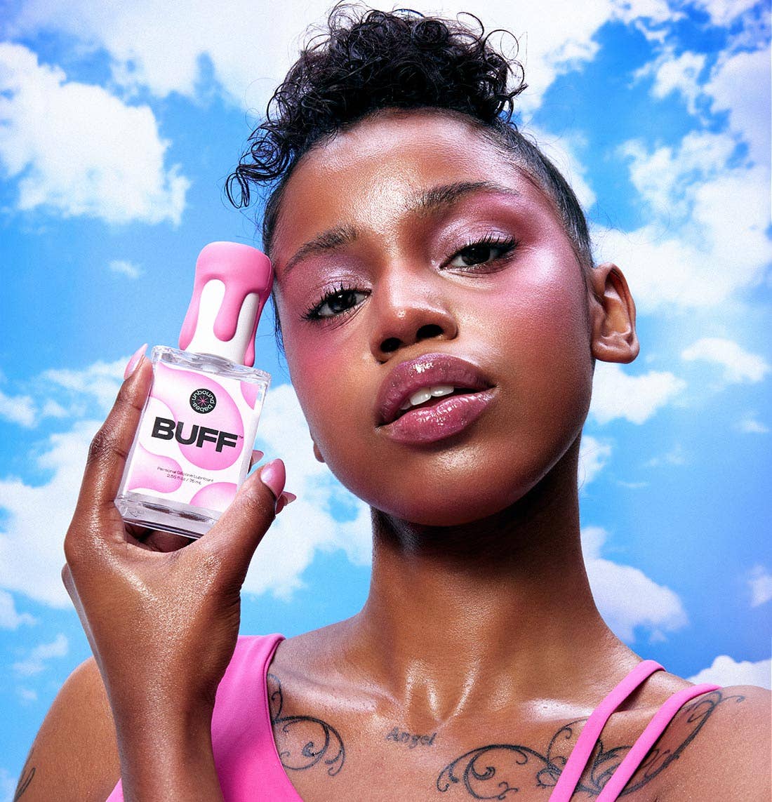 A woman holding a pink and clear bottle of Buff athletic cream against a bright blue sky with clouds background.
