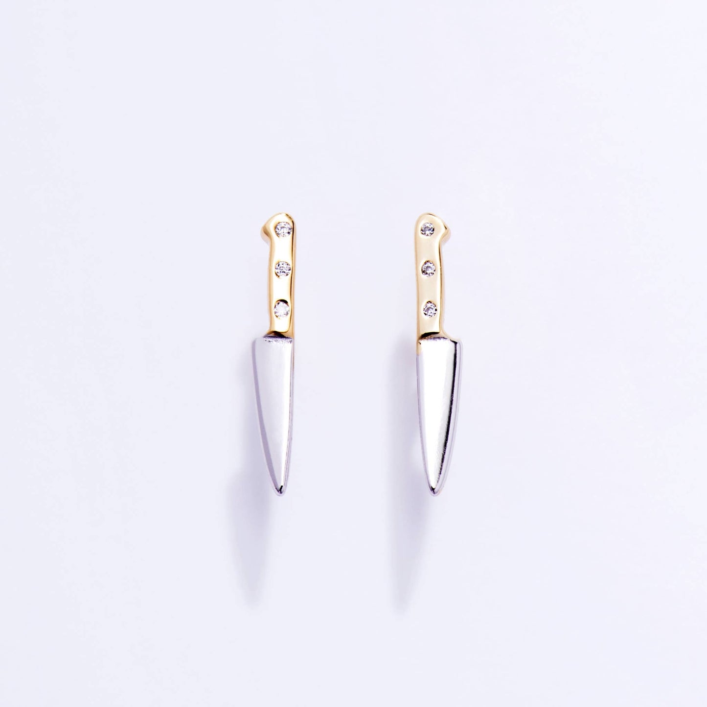 Pair of knife-shaped earrings with gold tops and silver blades on a light purple background