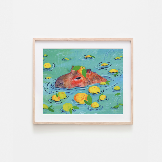 A framed art print featuring a whimsical illustration of a capybara swimming in turquoise water with yellow lemons and green leaves, set against a clean white background.