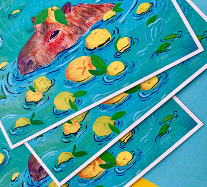 Three overlapping art prints featuring a whimsical illustration of a hippopotamus in turquoise water with floating yellow lemons, displayed on a blue surface.