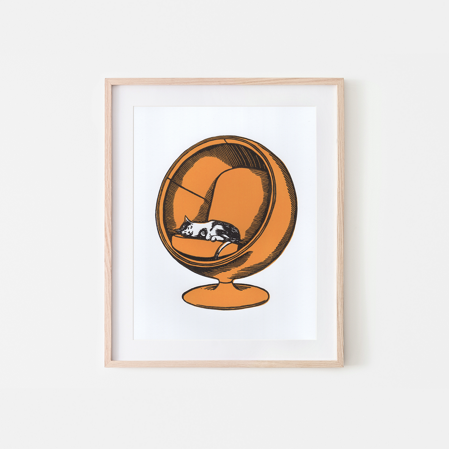 Framed wall art print featuring a stylized orange spherical chair with a sleeping black-and-white cat inside, shown in a light wood frame against a white background.