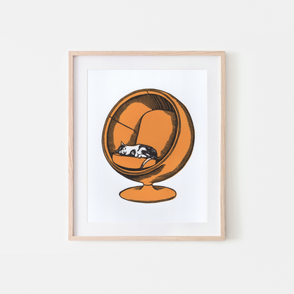 Framed wall art print featuring a stylized orange spherical chair with a sleeping black-and-white cat inside, shown in a light wood frame against a white background.