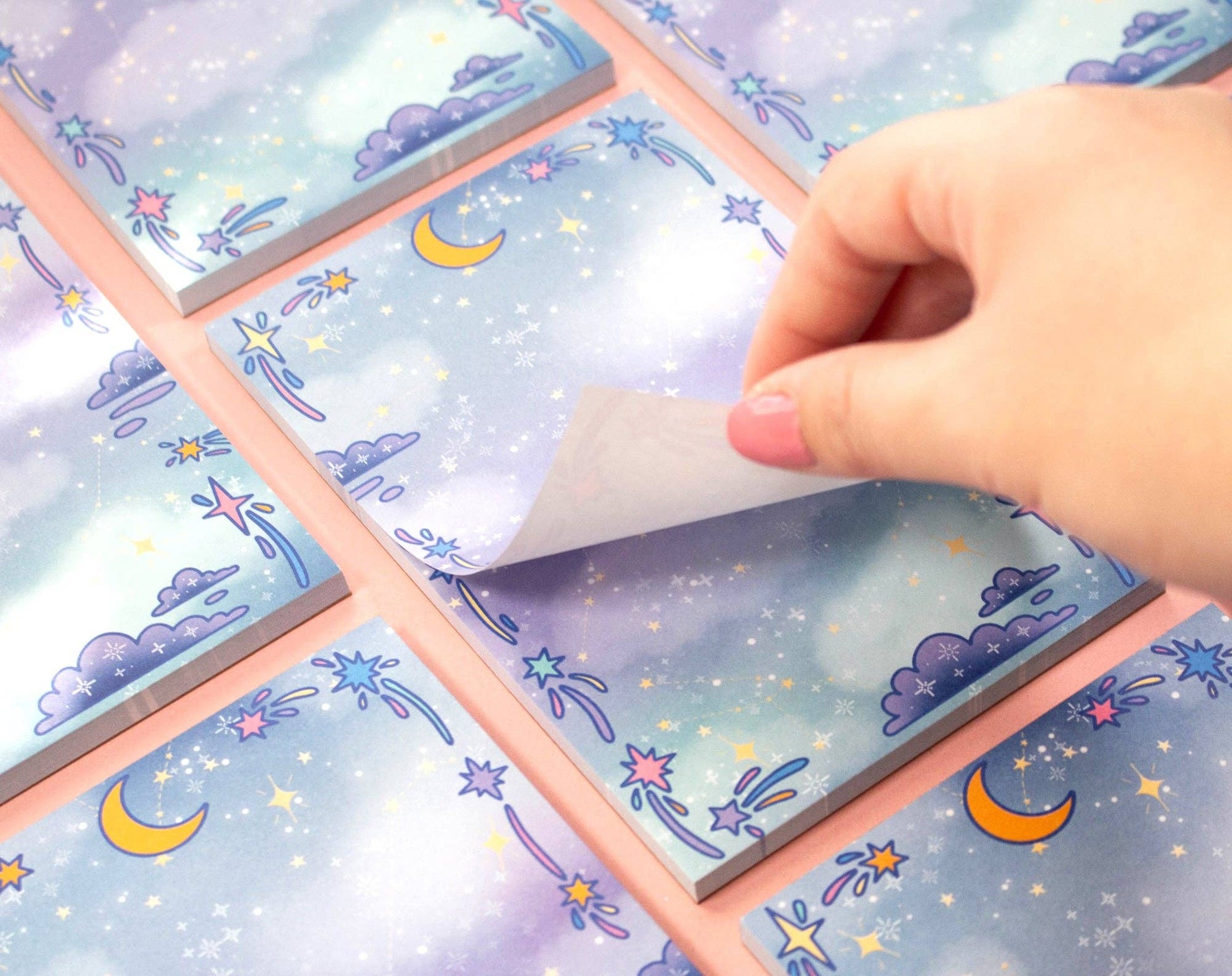 Hand lifting a page from a notepad with celestial moon and star design on a surface.