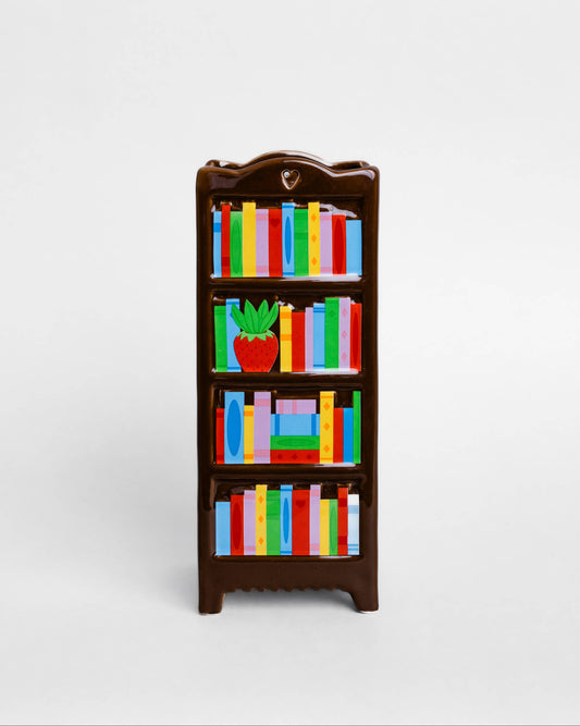 A miniature dark brown bookshelf vase with colorful books and a small potted plant, centered on a light gray background.