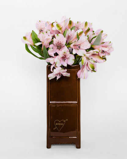 A dark brown wooden pedestal vase with a heart and arrow engraving, filled with pink alstroemeria flowers, set against a plain white background.