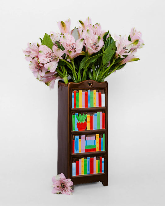 A decorative dark brown bookshelf-shaped vase filled with pink alstroemeria flowers and green leaves, set against a plain light gray background.