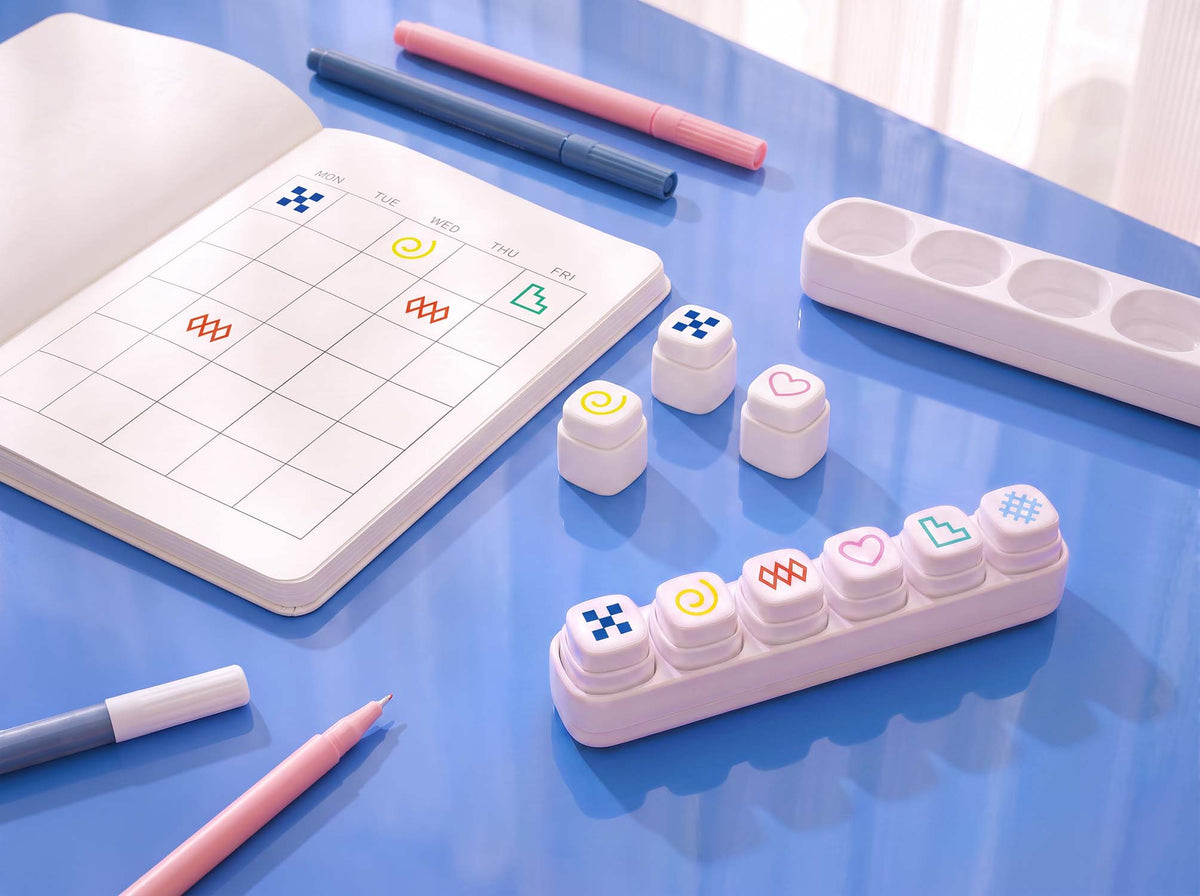 A pastel desk organizer set arranged on a blue tabletop, including a white grid planner pad, pink and blue pens, a white tray, and a row of white decorative button-like pieces with colorful symbols.