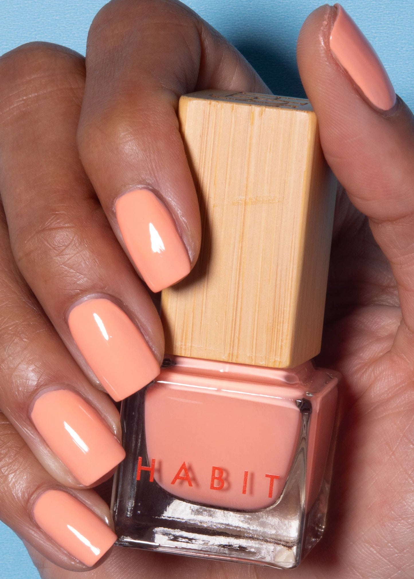 Hand holding a bottle of Habit nail polish with peach-colored nails against a blue background