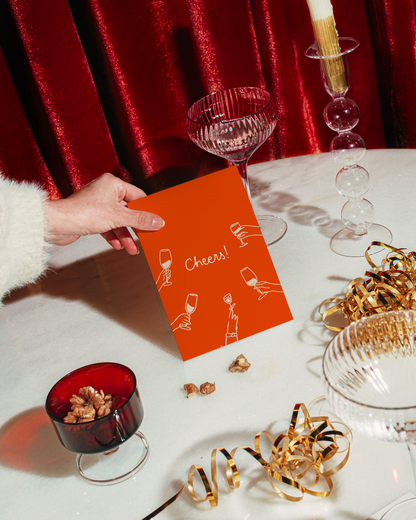 A hand in a white sleeve holds an orange greeting card that says 'Cheers!' with simple white illustrations. The card is shown in a festive tabletop setting with red velvet curtains, gold ribbons, and glassware.