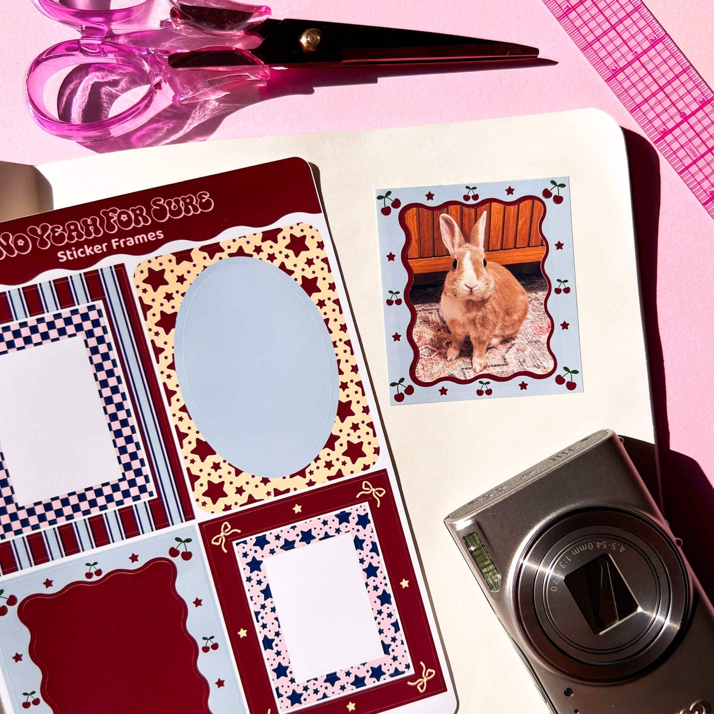 A styled flat-lay of a sticker frame sticker sheet and a sample decorative sticker featuring a brown rabbit on a pink background, arranged with scissors, a camera, and a pink ruler on a light surface.