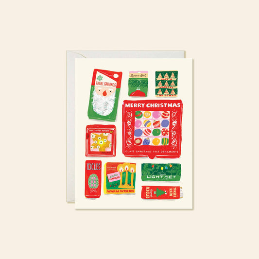 A square Christmas greeting card with a white envelope behind it, featuring a collage of vintage-style holiday illustrations and text including 'MERRY CHRISTMAS' and 'GLASS ORNAMENTS.' The card is photographed on a plain warm beige background.
