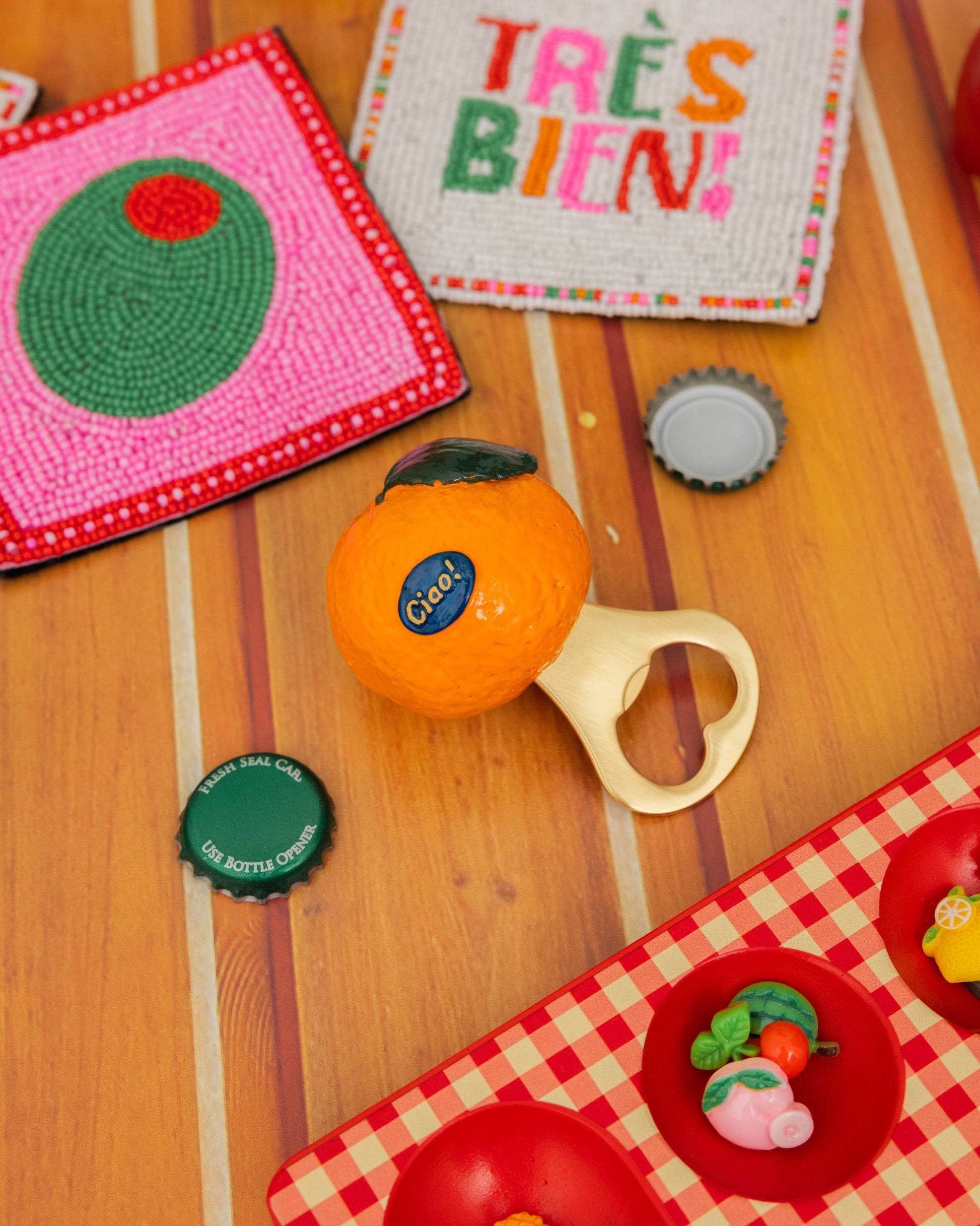 A styled flat-lay of handmade crocheted or knit accessories on a wooden surface, including a pink and green coaster, a white woven mat with colorful 'TRÈS BIEN!' text, an orange bottle opener, bottle caps, and a red-and-white check picnic mat.