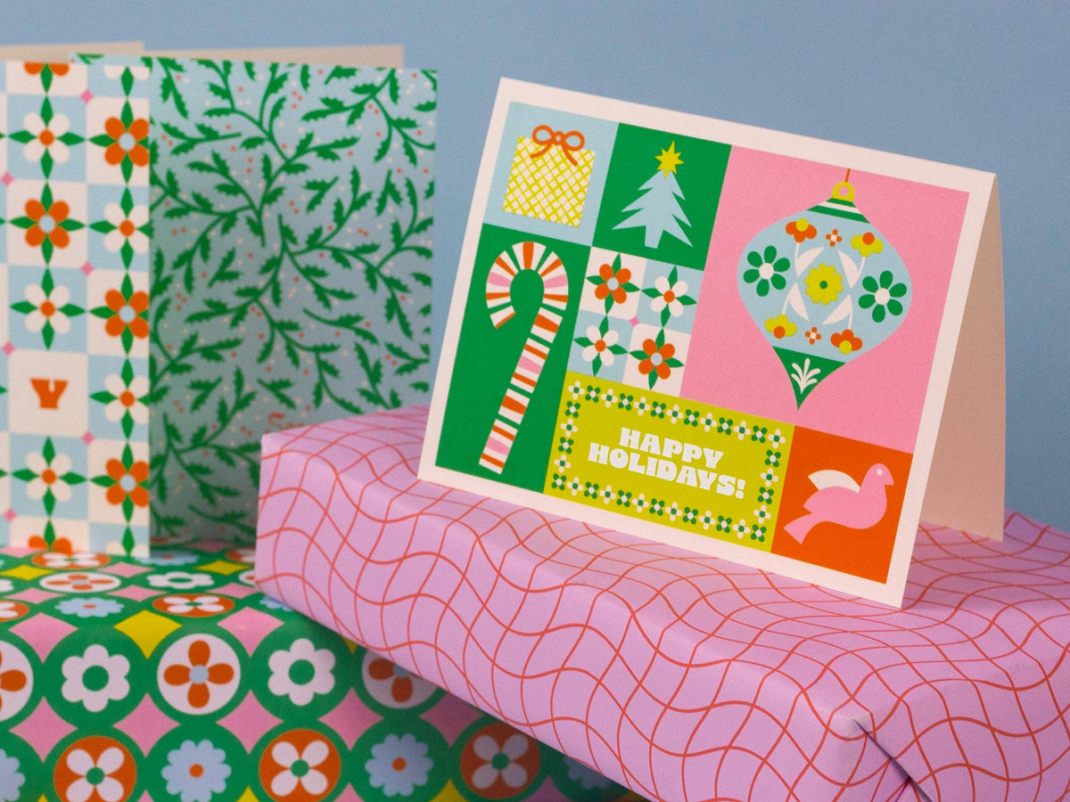 A festive holiday greeting card with retro 1960s-style patterns, shown standing on top of wrapped gift boxes. The card features a pink background with green, orange, yellow, and white holiday illustrations including a candy cane, ornament, Christmas tree, and the text 'HAPPY HOLIDAYS!'.