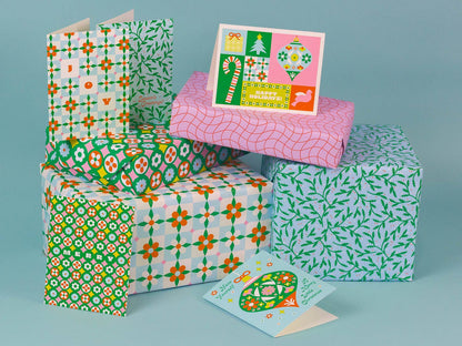 A collection of Christmas-themed gift boxes wrapped in colorful patterned paper, with two matching greeting cards displayed in front, set against a solid light blue background.