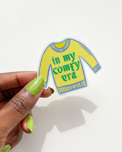 Sticker of a yellow sweater with text held by a hand on a white background, Comfy Era Sweater Sticker