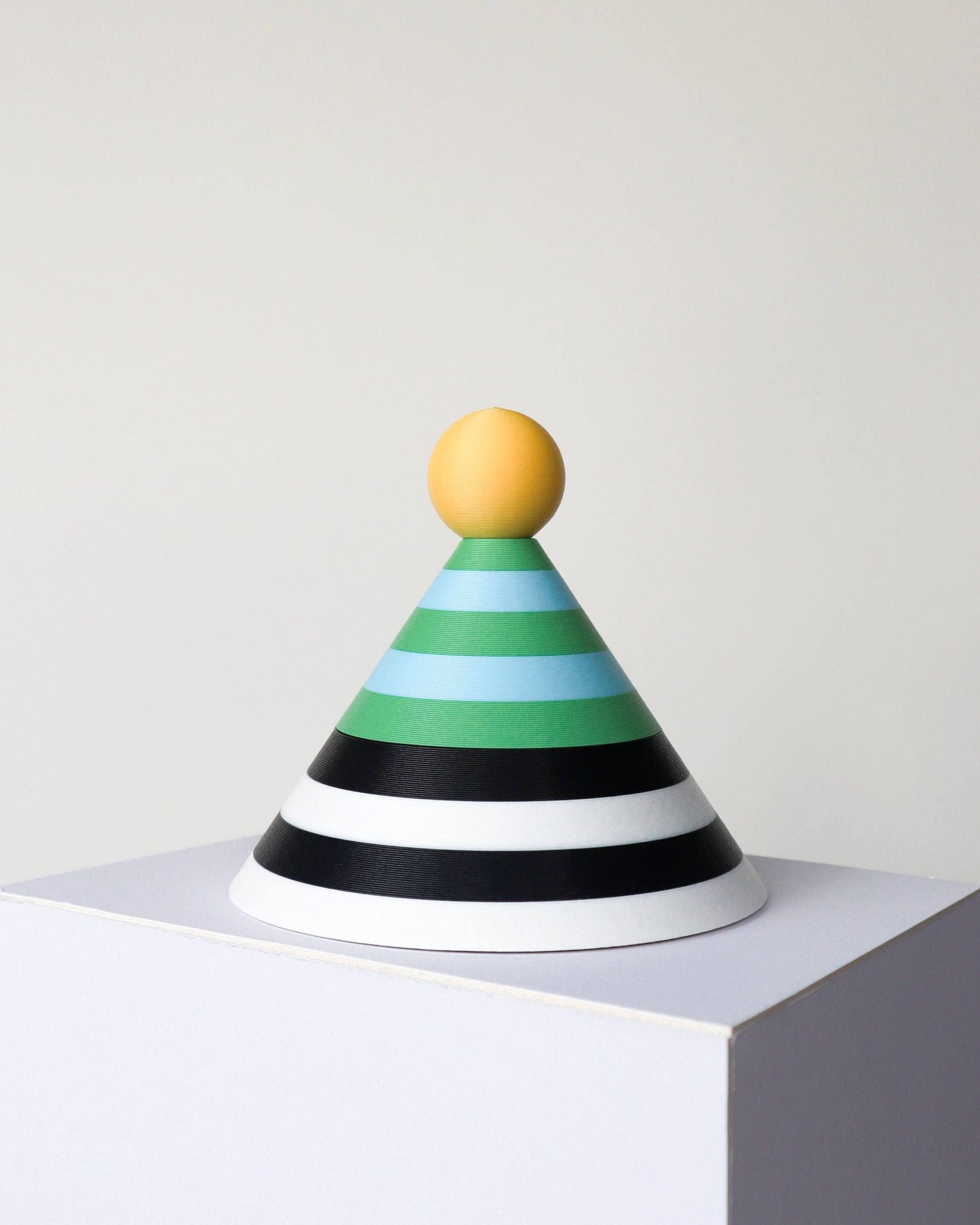 A cone-shaped dish with horizontal stripes in yellow, green, light blue, black, and white, topped with a round yellow handle, displayed on a white pedestal against a light gray background.