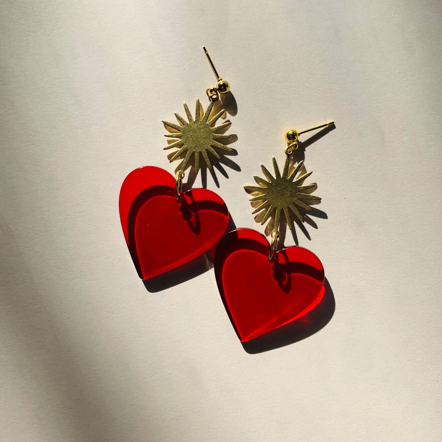 A pair of gold-tone dangle earrings with sunburst tops and translucent red heart-shaped pendants, photographed on a light neutral background with strong directional shadows.