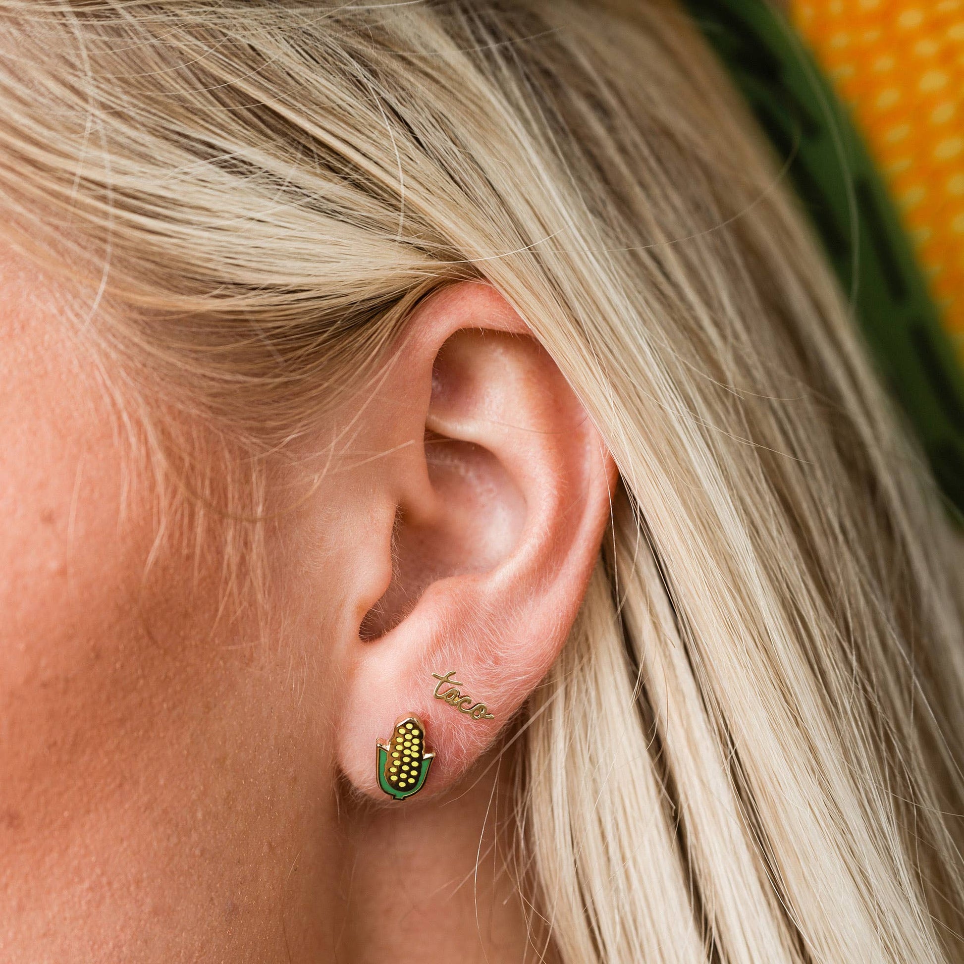 Close-up of an ear wearing a gold earring with green accents and 'tacos' , Corn on the Cob Enamel Stud Earrings Larissa Lodenengraved.