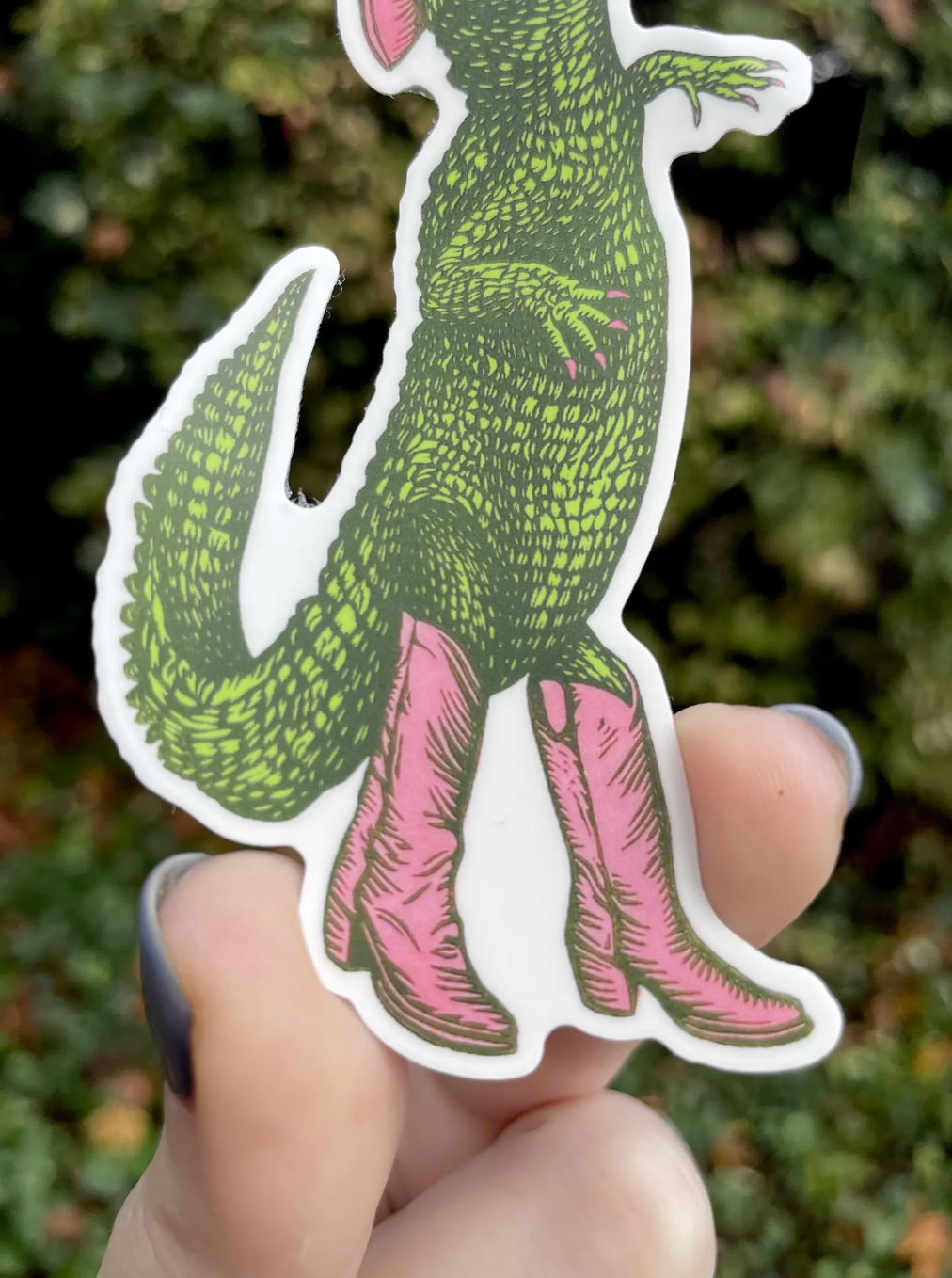 Sticker of a green creature with pink boots held by a hand against a blurred natural background