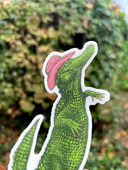 Sticker of a green alligator with a pink bow on a blurred green background