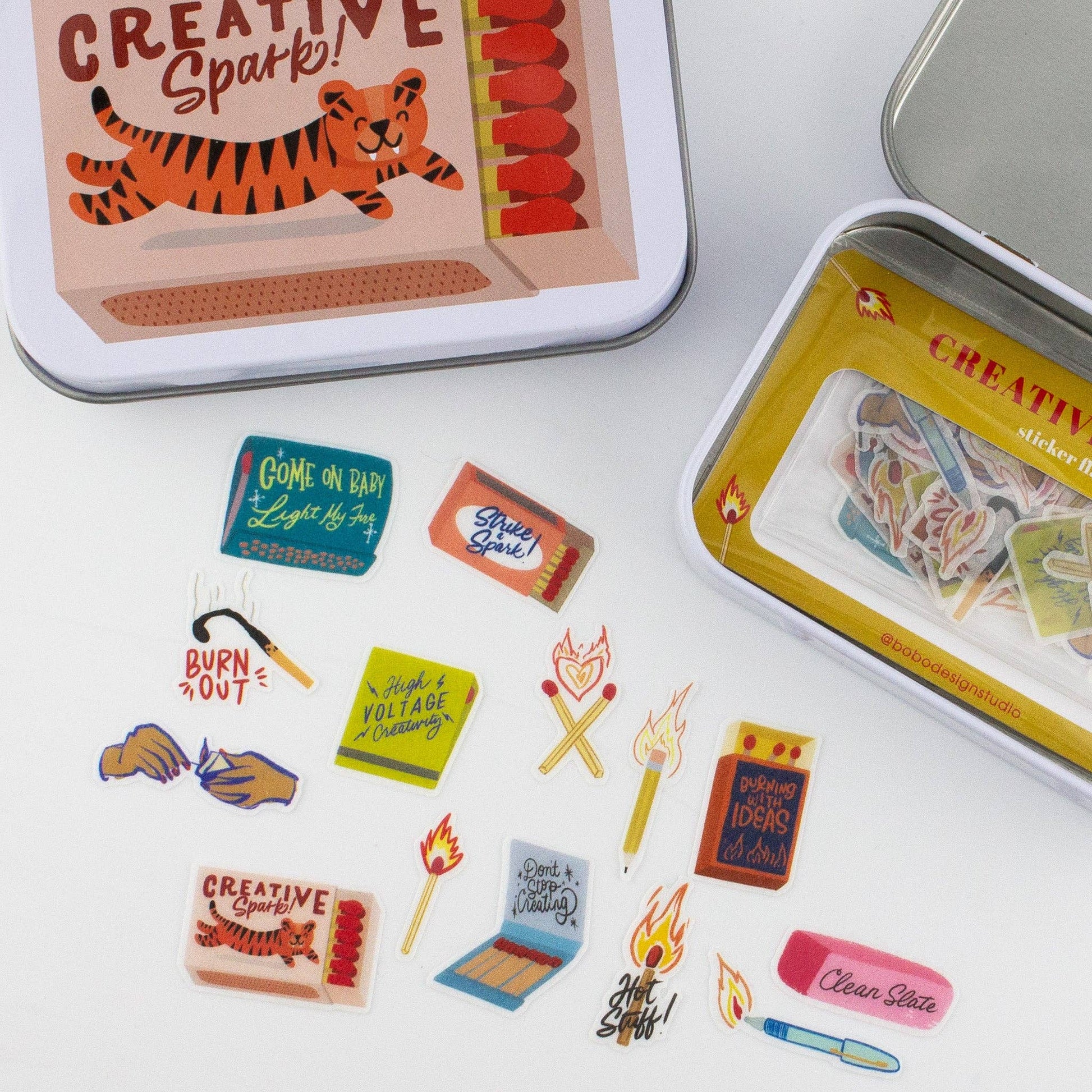 A flat-lay display of various small decorative stickers inspired by matchboxes and fire symbols, shown alongside a metal tin of matching stickers on a white background.